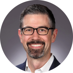 Watt Hamlett, smiling against a gray background. He has dark hair, a beard, and glasses, and is wearing a white shirt and dark jacket.